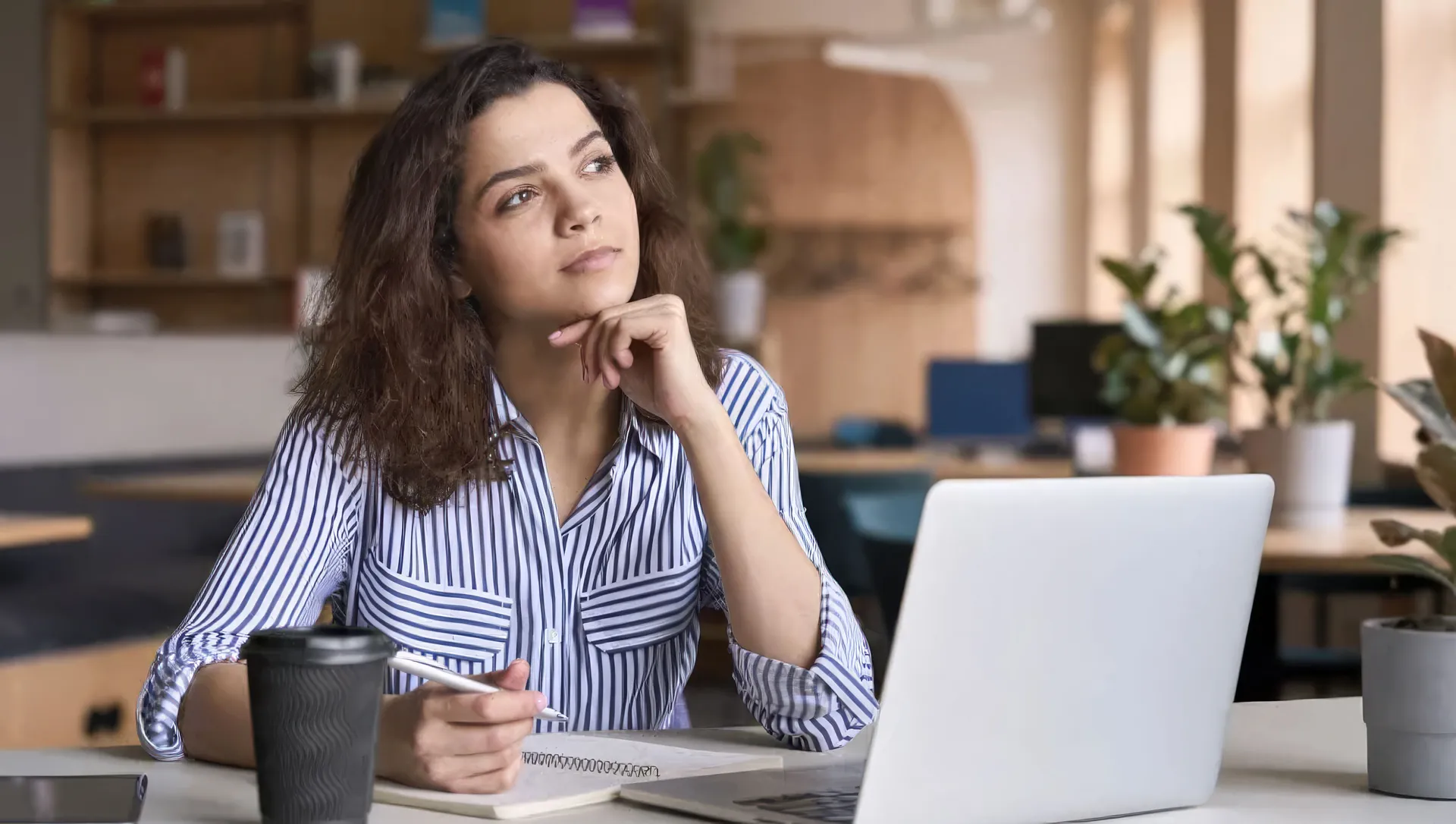 A professional pauses to reflect while taking notes beside her laptop in a bright, plant-filled workspace